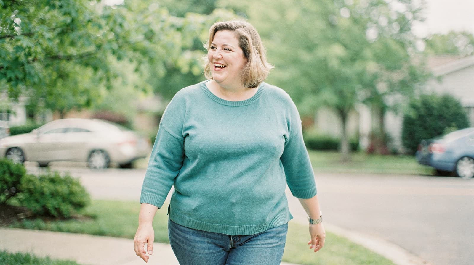 A woman in a teal sweater walks and smiles on a suburban street with trees and parked cars in the background.