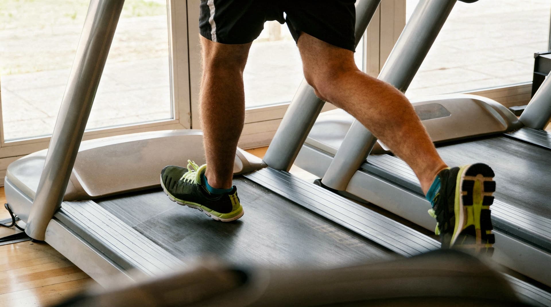A person in athletic shoes runs on a treadmill indoors near large windows.