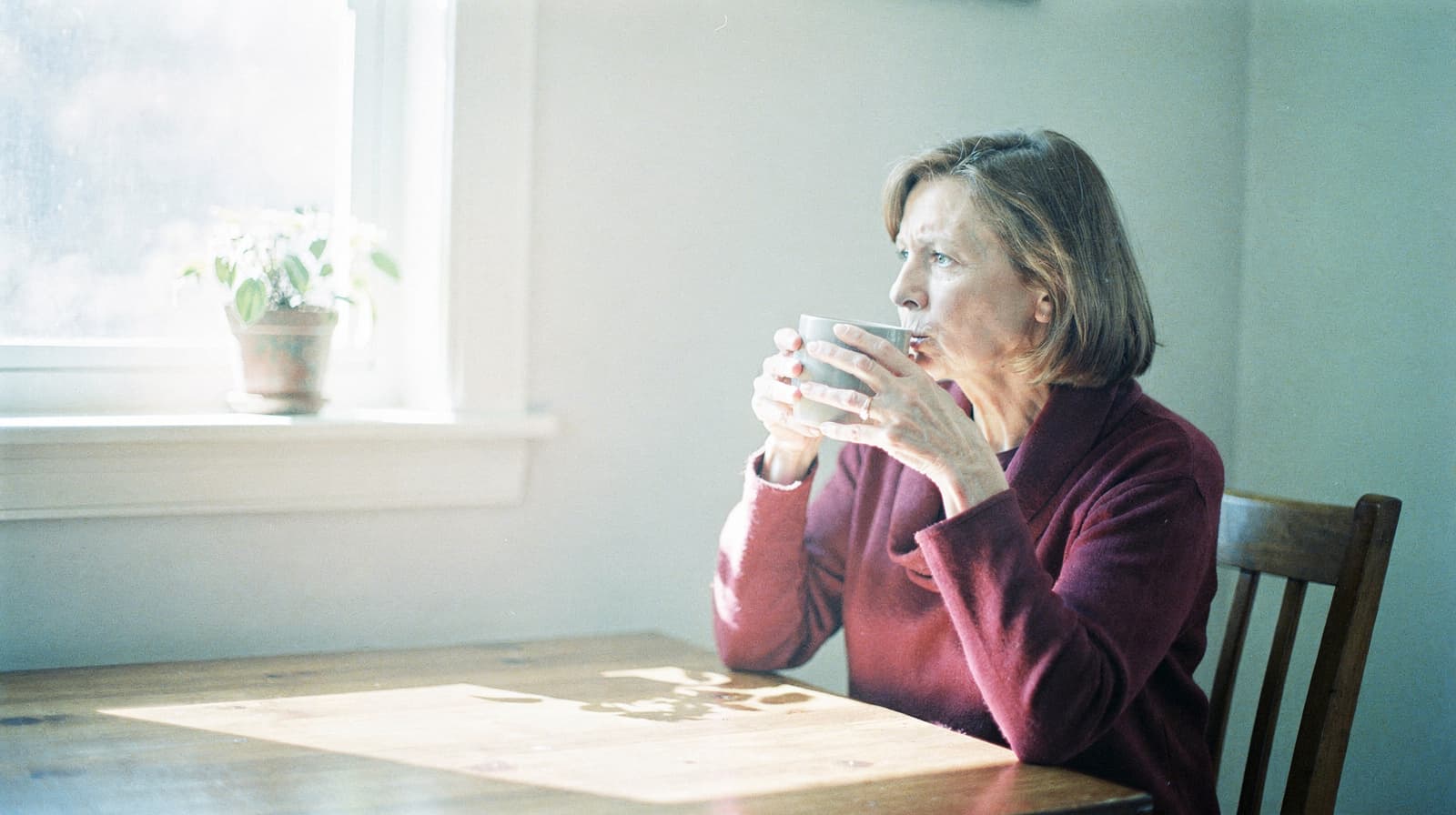 A woman in a red sweater sits at a wooden table, drinking from a mug and looking out a sunlit window with a potted plant.
