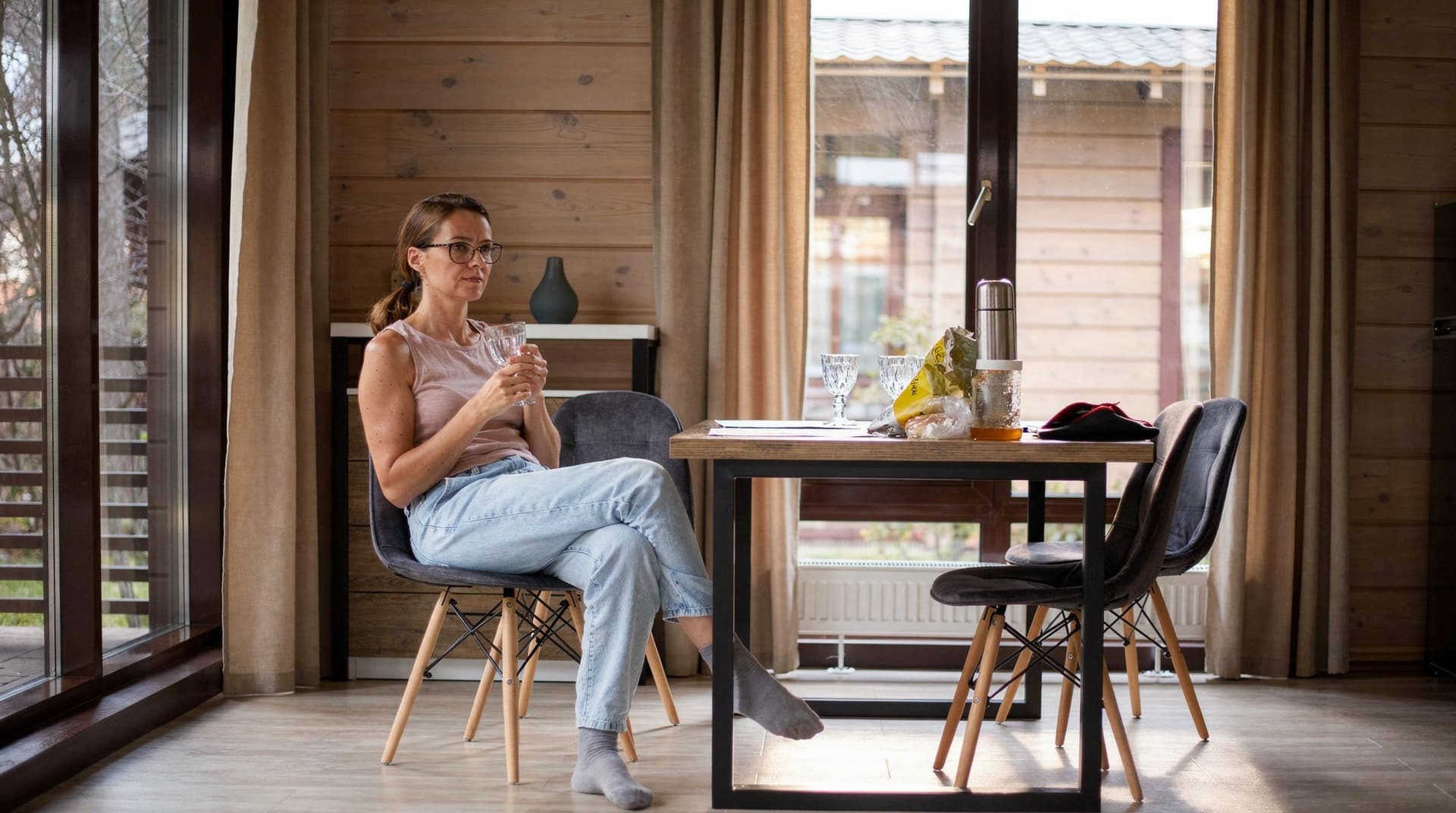 A woman sits at a dining table in a wooden room, holding a glass and looking out the window.