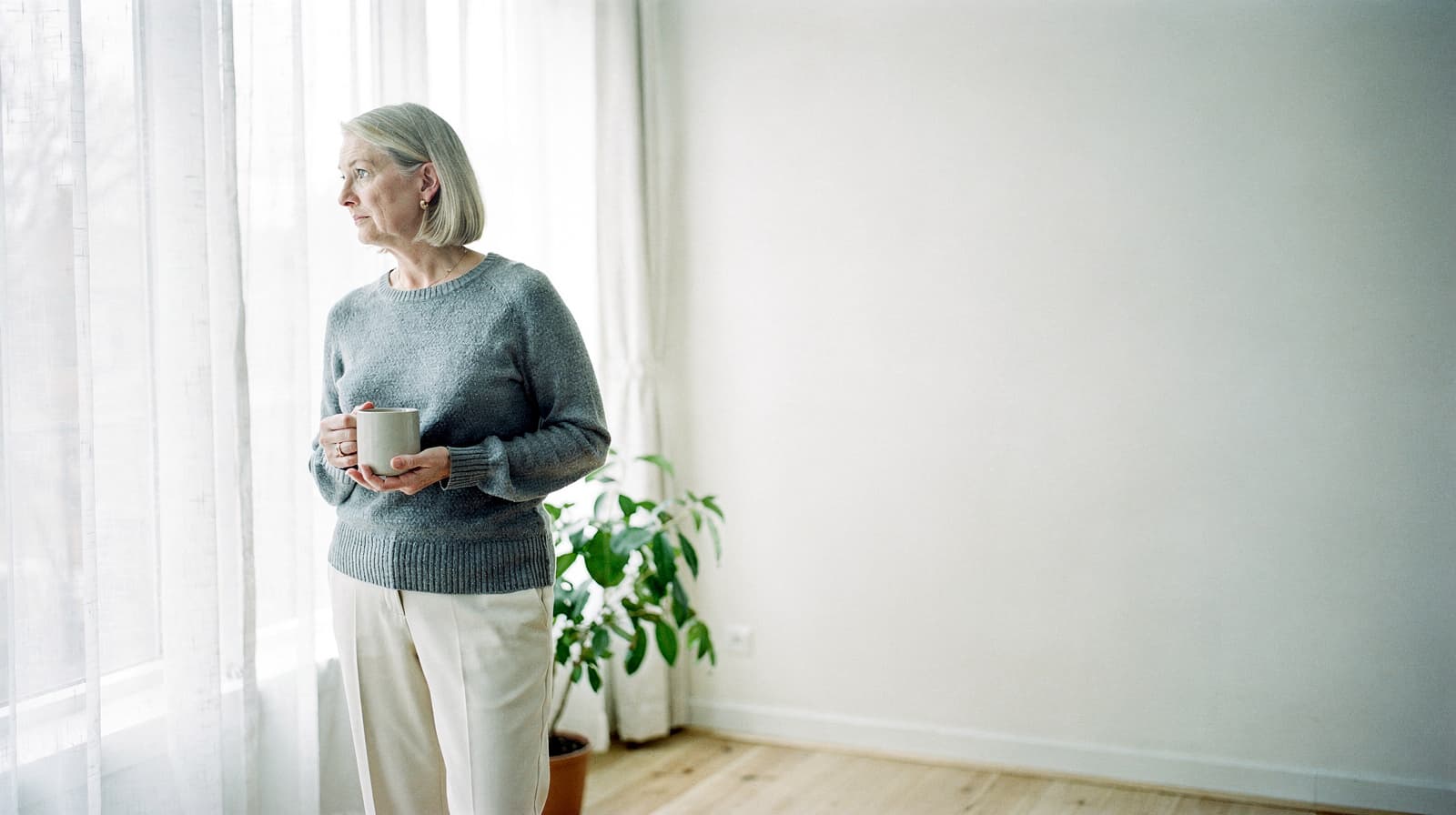 A woman holding a mug looks out a window in a bright room with a potted plant.