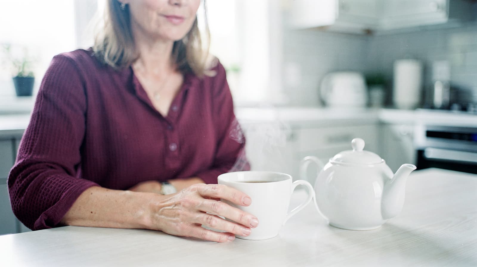 A woman in a red shirt sits at a kitchen table holding a steaming cup next to a white teapot.
