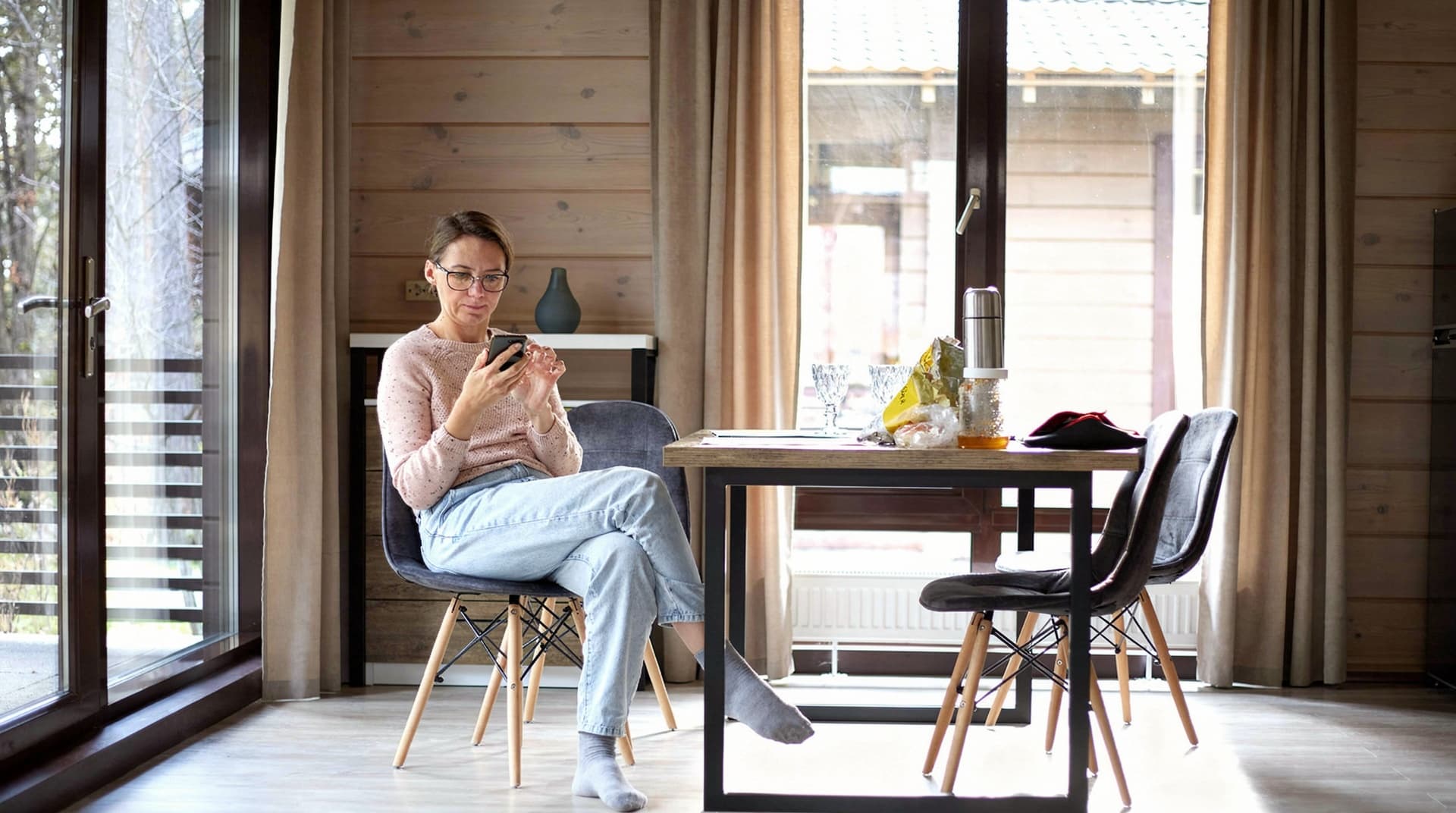 A woman sits at a dining table in a wooden room, looking at her phone with snacks and drinks on the table.
