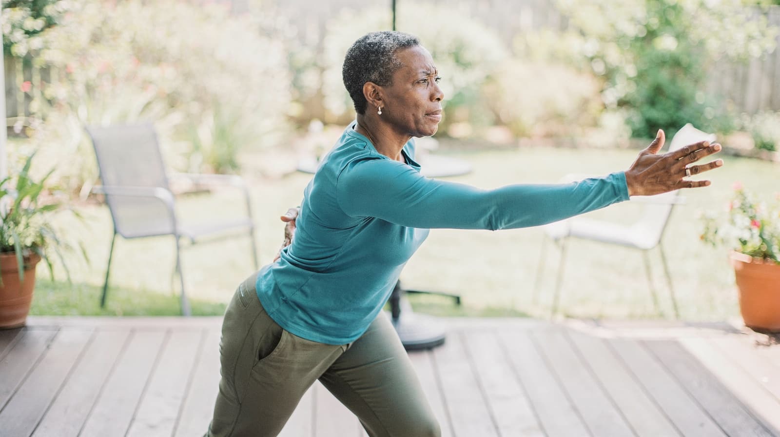 A woman in a teal shirt stretches her arm forward while exercising on a wooden deck in a garden.