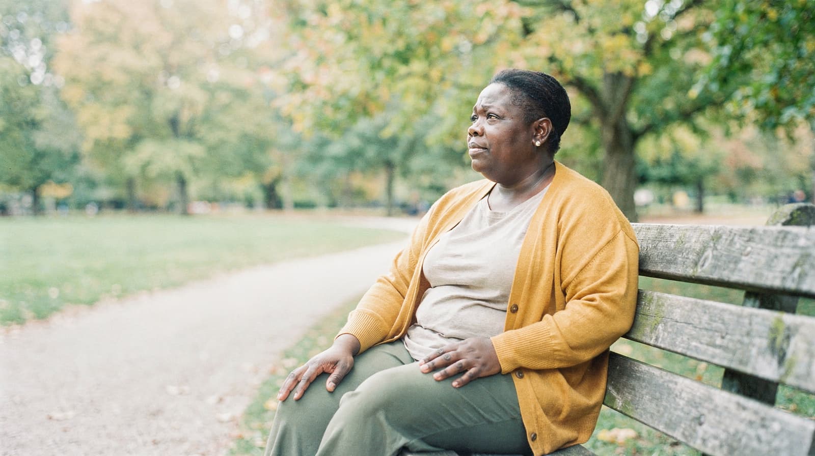 A woman sits on a park bench looking into the distance, surrounded by trees and a gravel path.