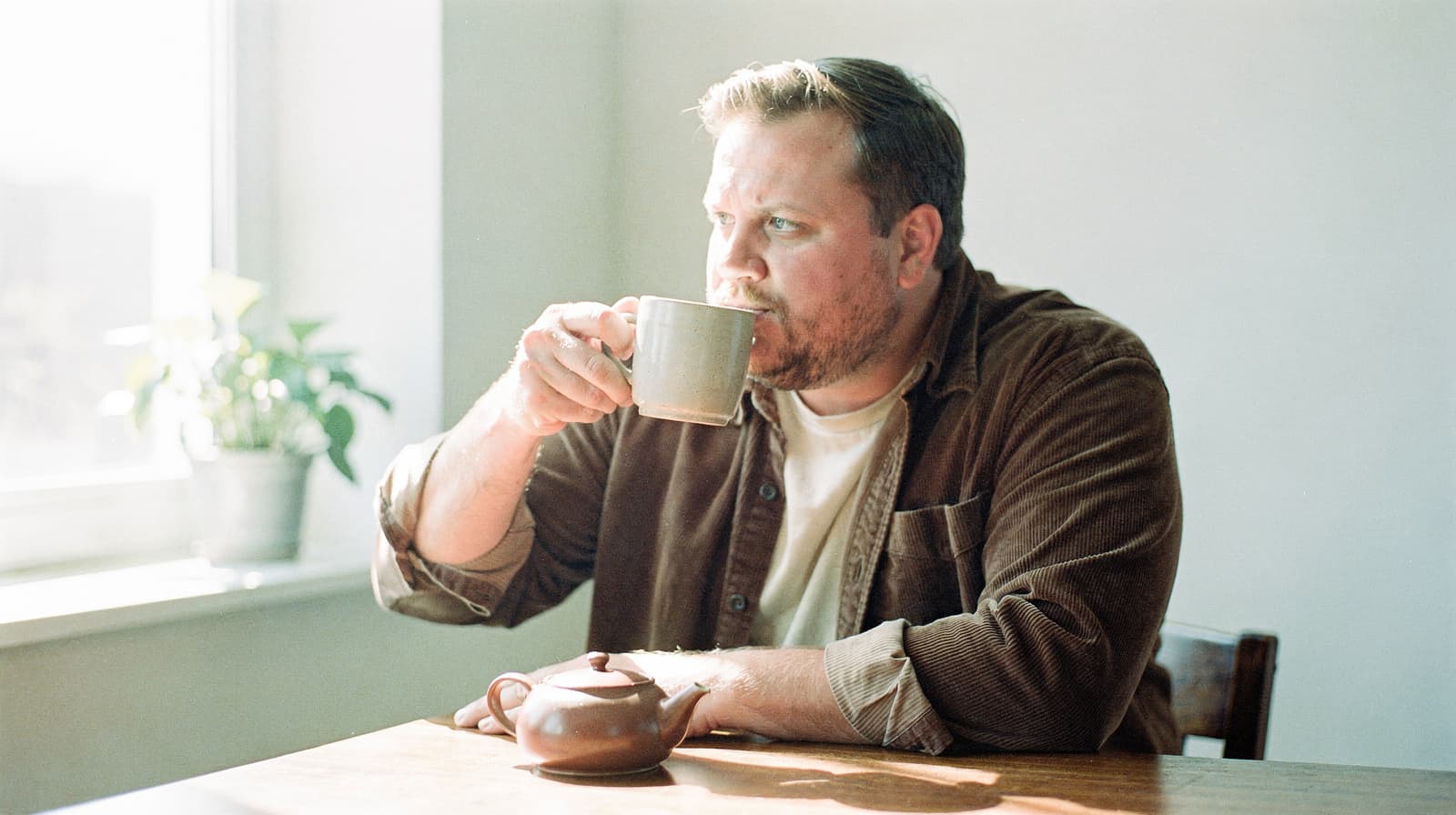A man sits at a table drinking from a mug with a teapot nearby, next to a window with a potted plant.