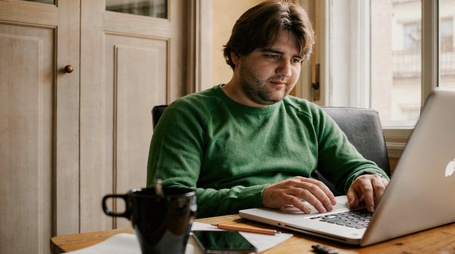 A man in a green sweater works on a laptop at a wooden table with a mug and smartphone nearby.