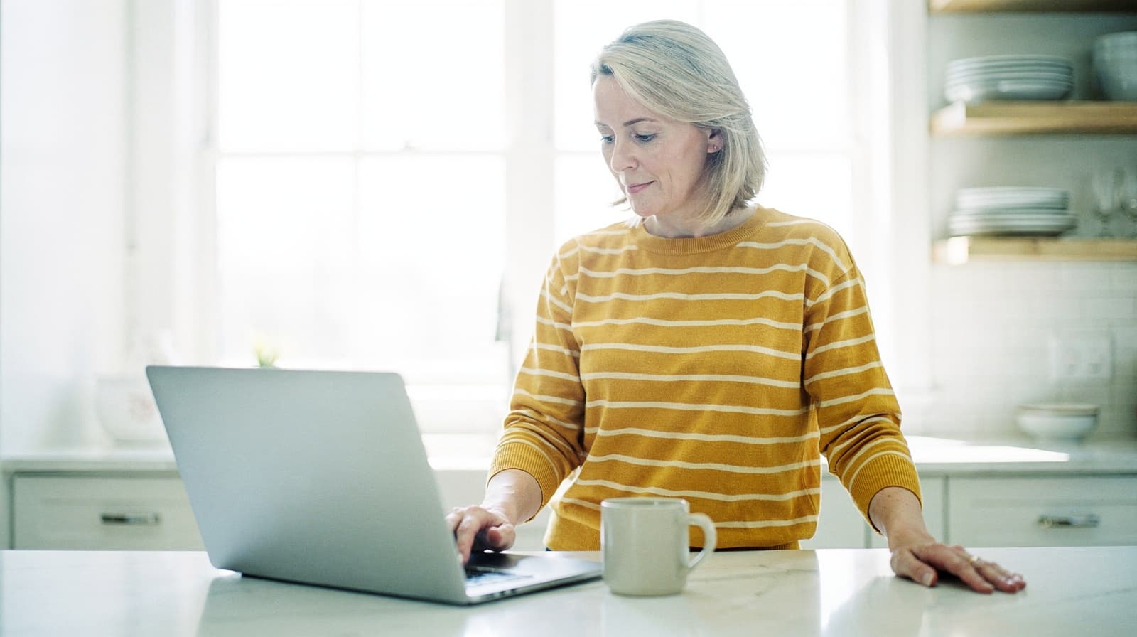 A woman in a striped sweater uses a laptop at a kitchen counter with a mug beside her.