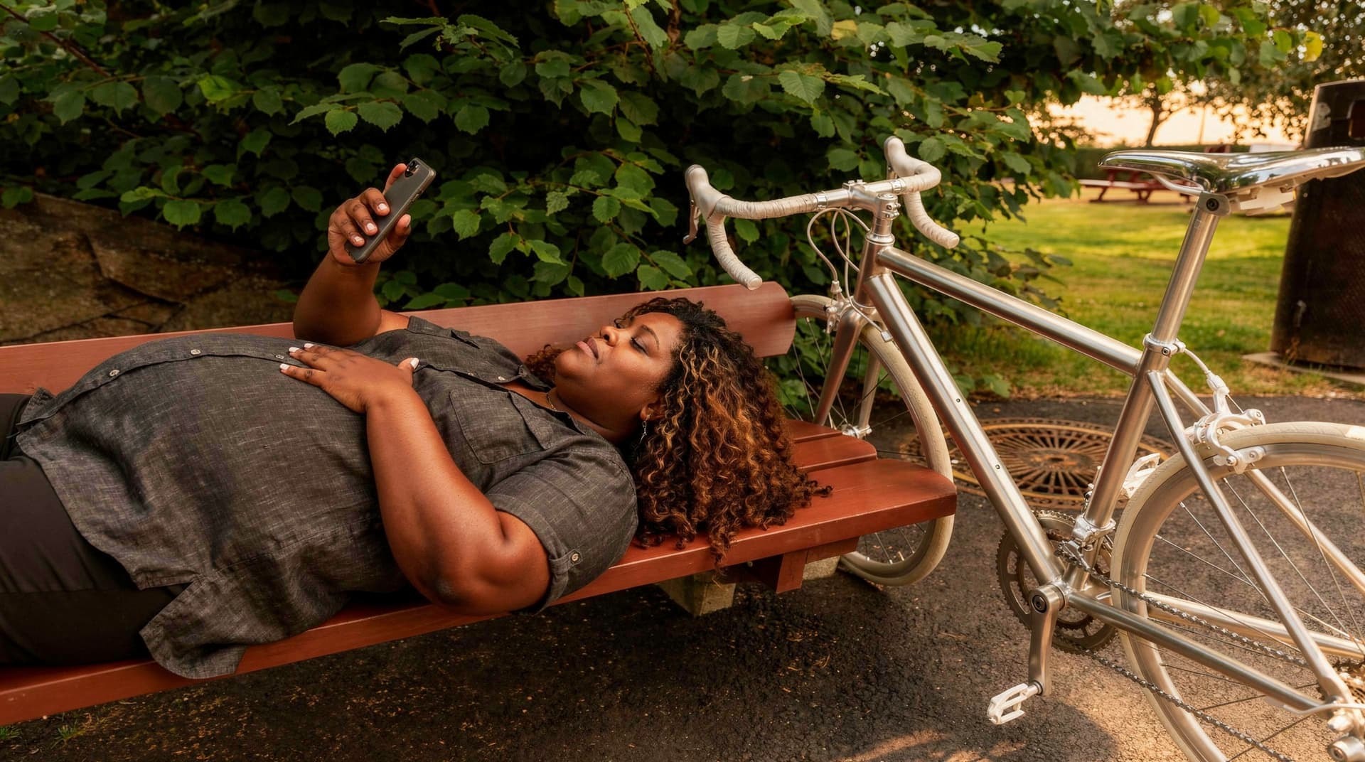 A woman lies on a park bench looking at her phone with a bicycle parked beside her.