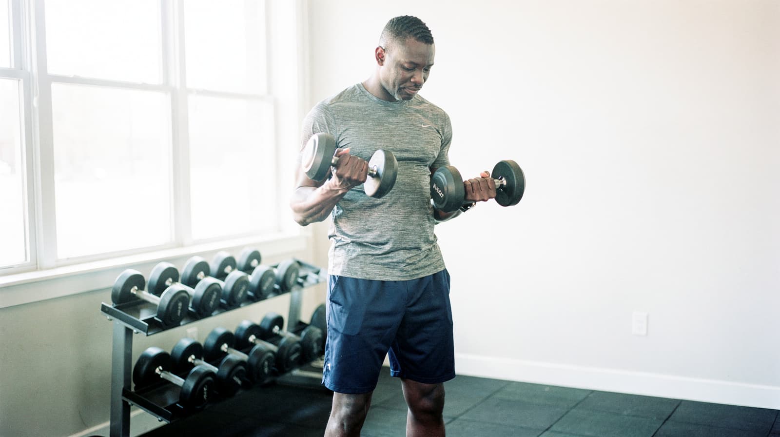 A man in a gym lifts dumbbells near a rack of weights by a large window.