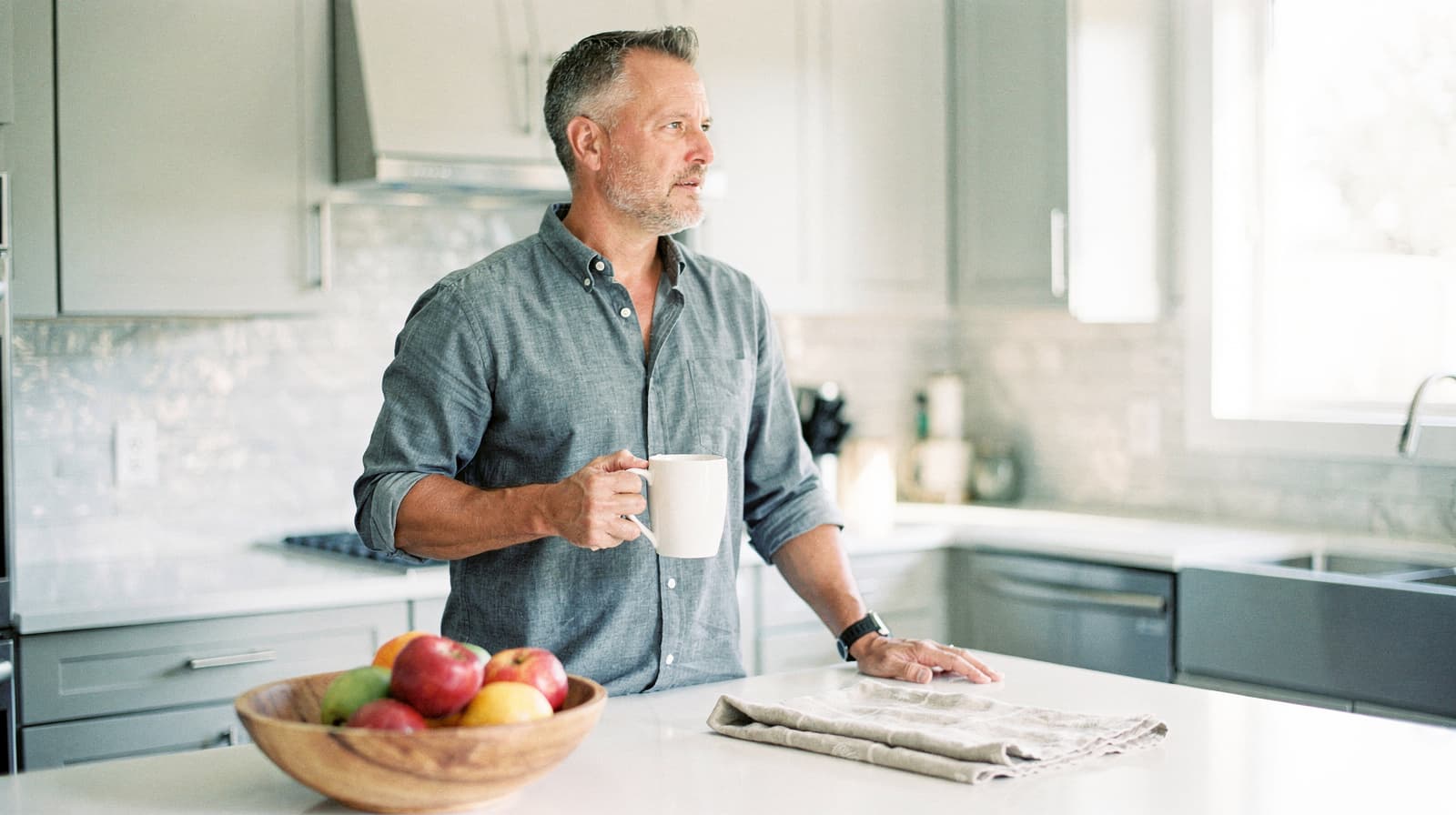 A man holding a mug stands in a kitchen beside a bowl of apples on a countertop.