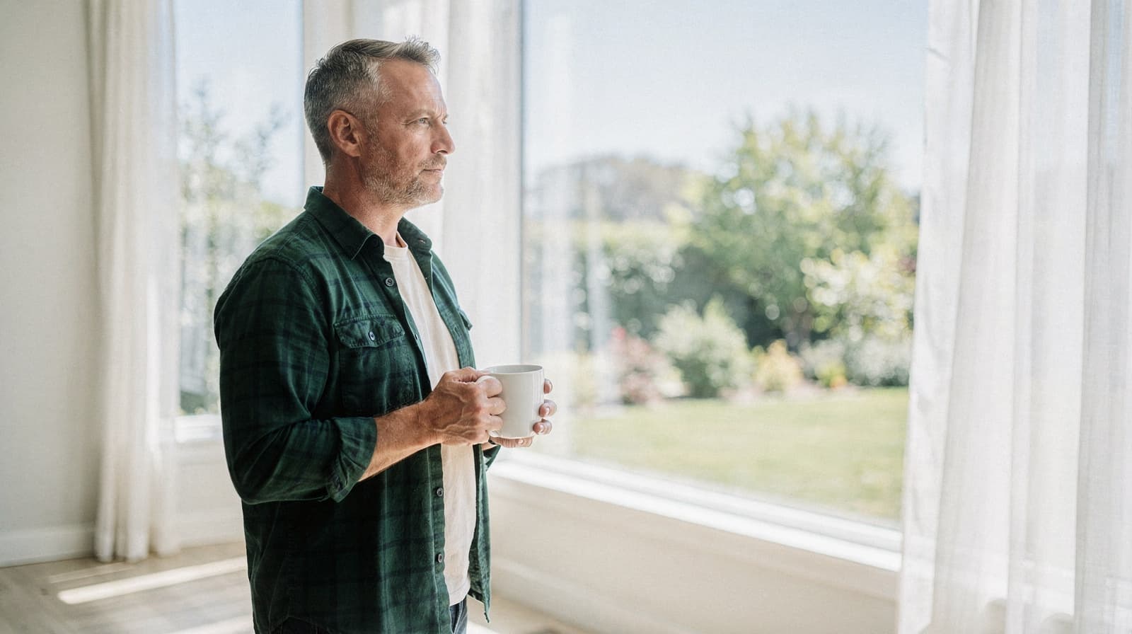 A man holding a mug looks out a window in a bright room with a view of a garden.