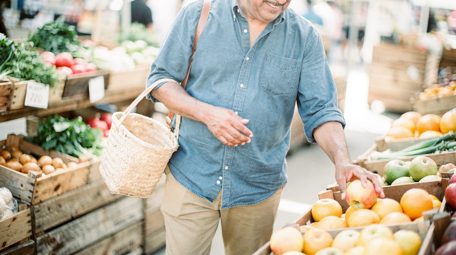 A man in a denim shirt selects fruit from a wooden crate at an outdoor market.