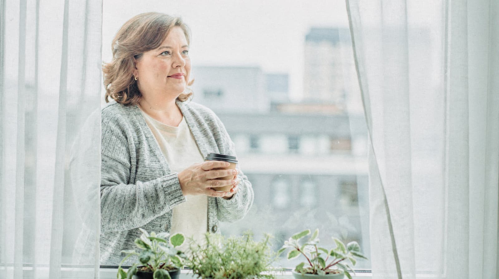 A woman holding a coffee cup stands by a window with plants on the sill, looking outside.