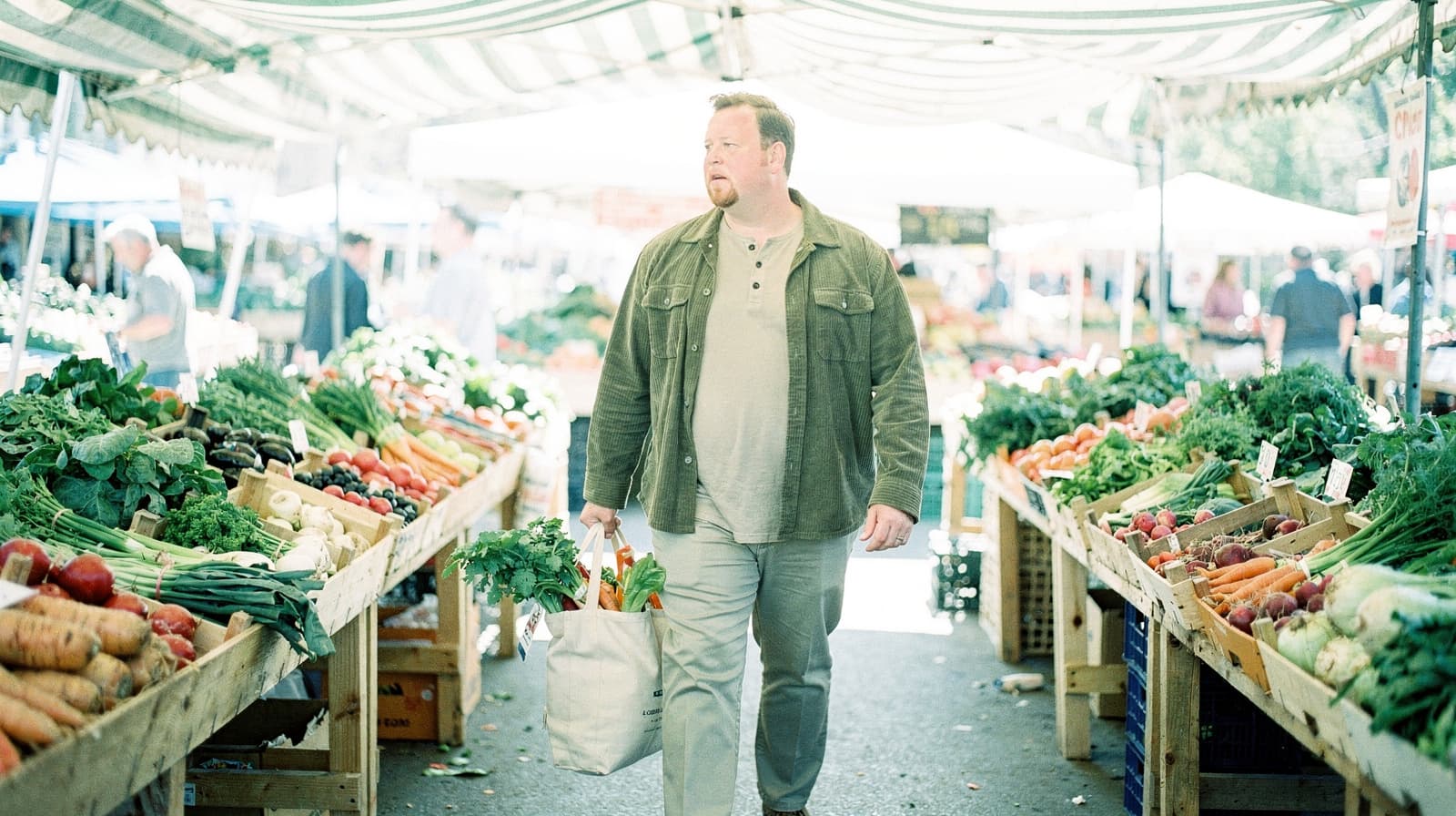 A man carries a bag of vegetables while walking through an outdoor market with various produce on display.