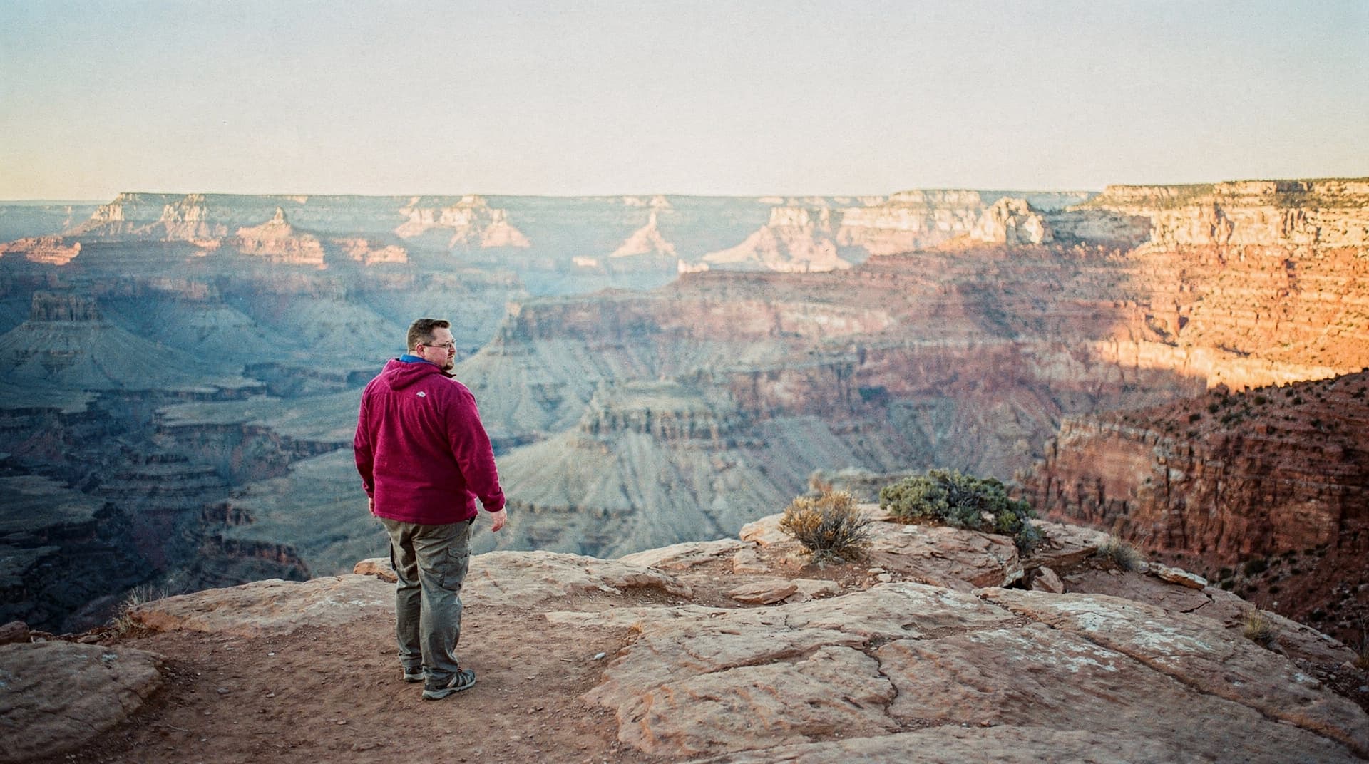 A person in a red jacket stands on a rocky cliff overlooking a vast canyon landscape.