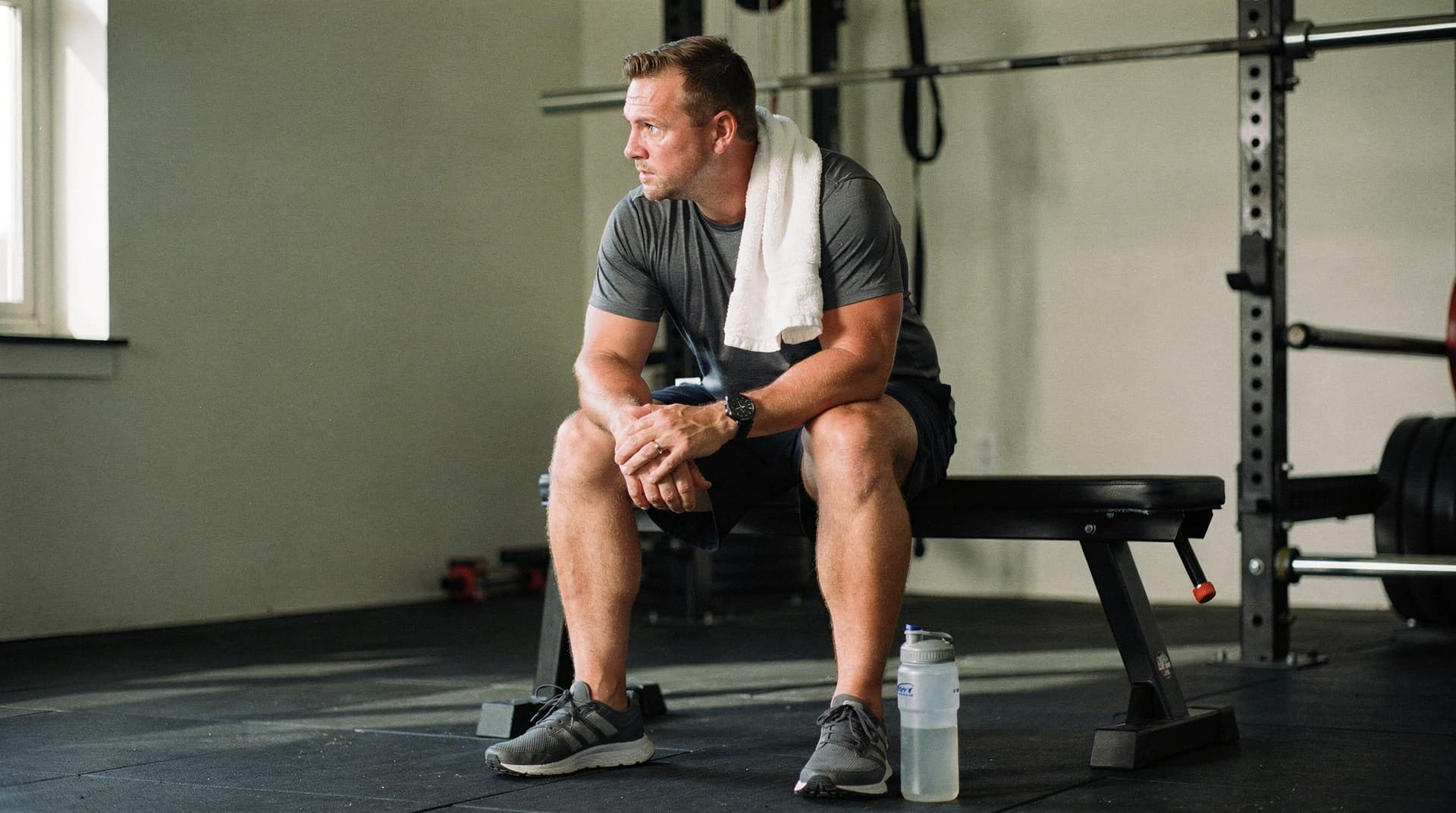 A man sits on a bench in a gym with a towel over his shoulder and a water bottle on the floor.