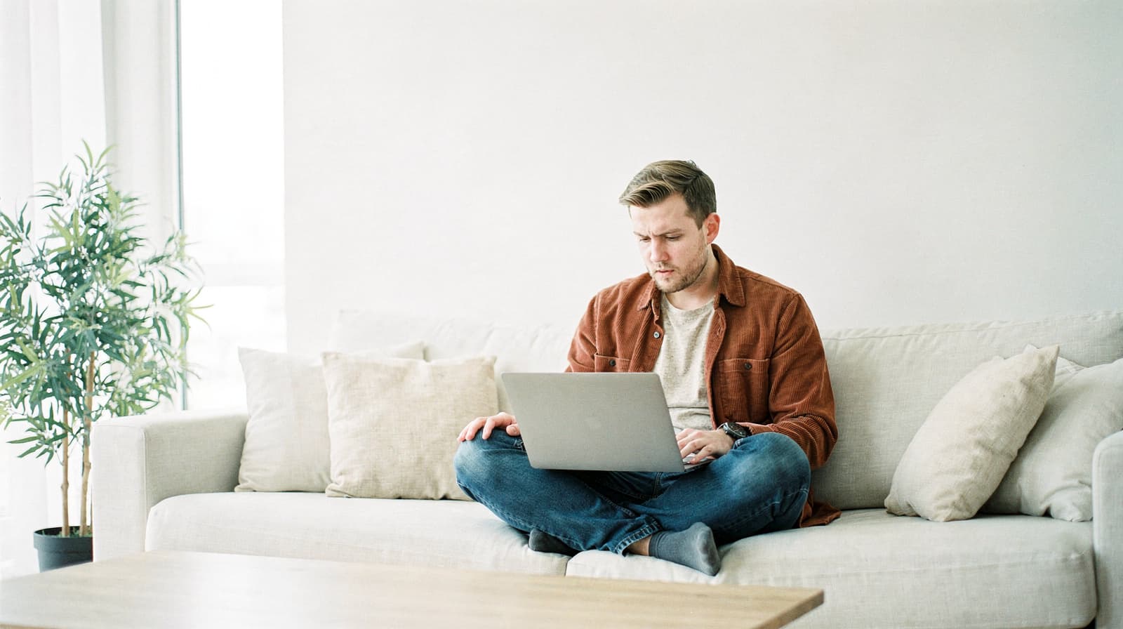 A man sits on a beige sofa using a laptop, with a plant and pillows in the background.