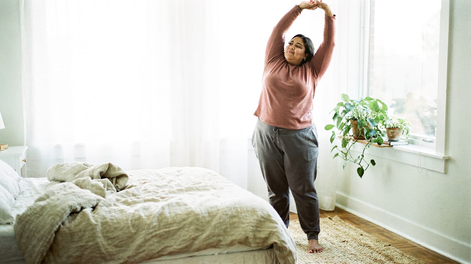 A woman stretches in a sunlit bedroom next to a bed and a window with plants.