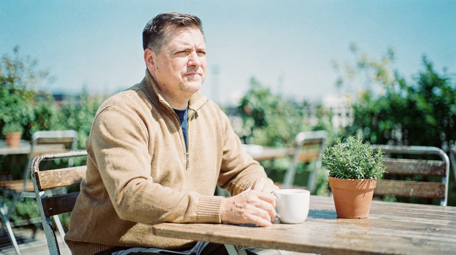 A man sits at an outdoor table holding a mug, with potted plants and empty chairs around him.
