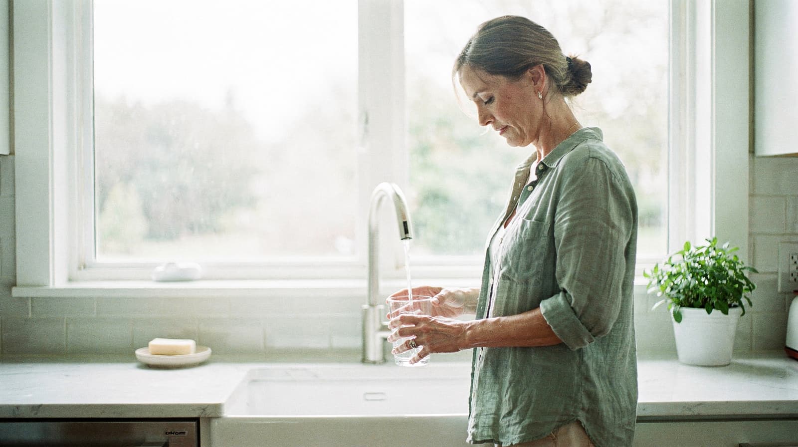 A woman stands at a kitchen sink filling a glass with water from the faucet.