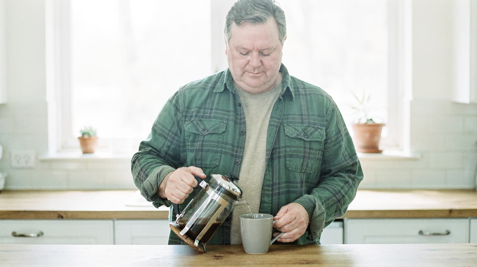 A man in a green shirt pours coffee from a French press into a mug in a bright kitchen.