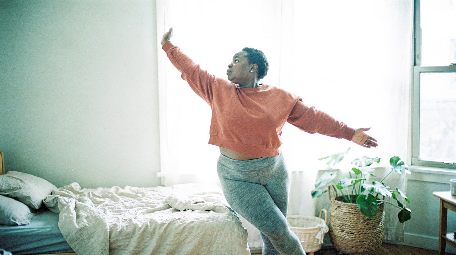 A person in a red sweater and gray pants stretches gracefully in a sunlit bedroom next to an unmade bed and a potted plant.