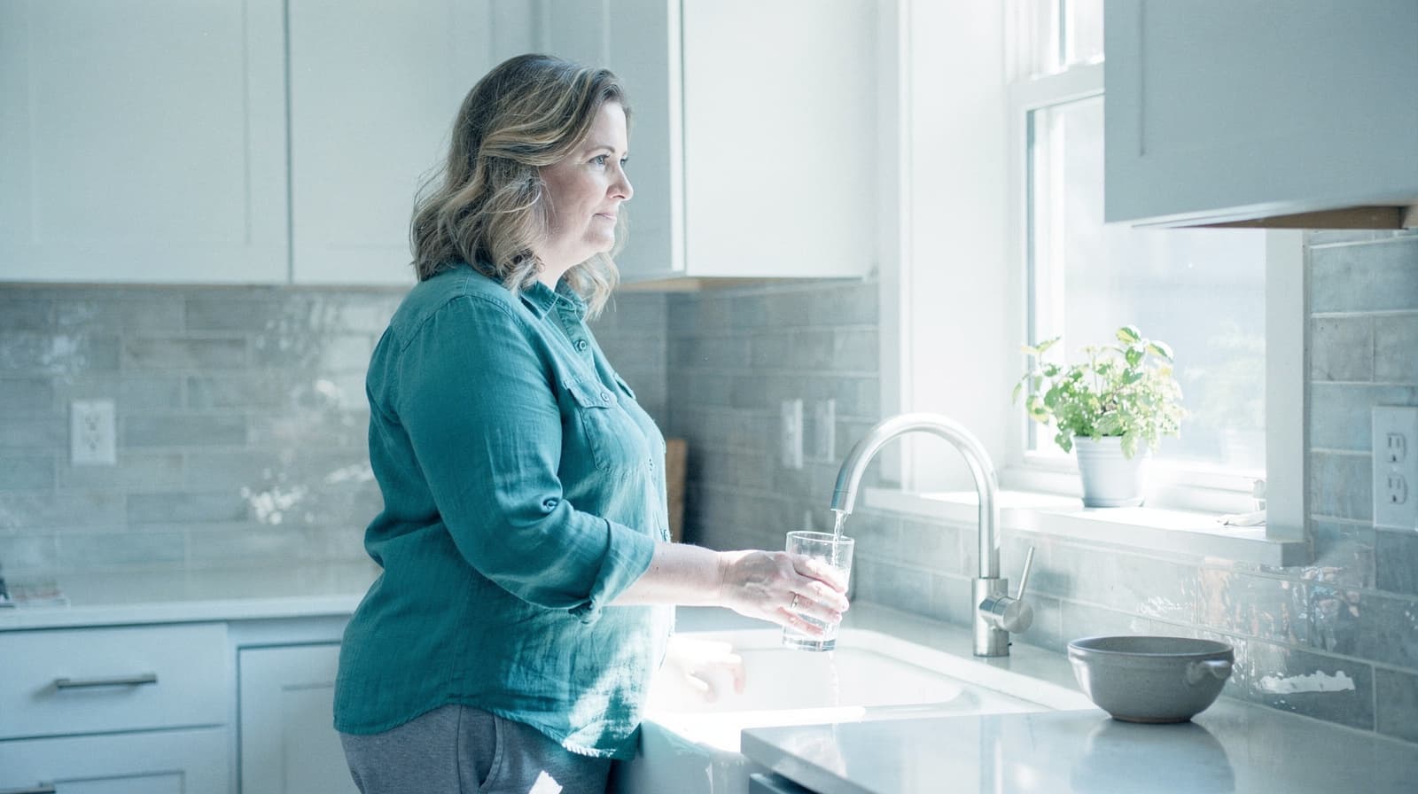 A woman stands in a kitchen by a window holding a glass of water.