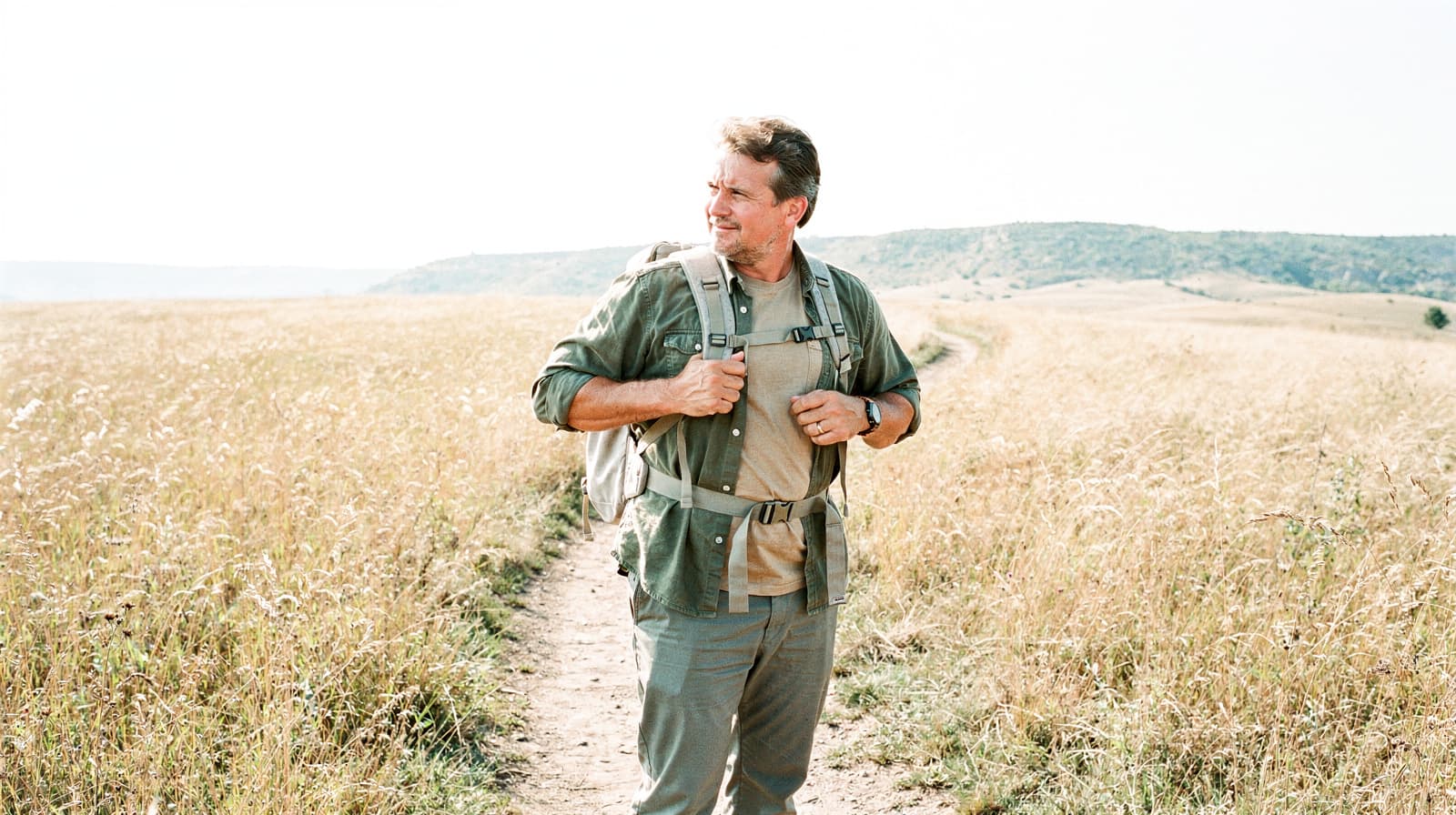 A man with a backpack stands on a dirt path in a grassy field under a clear sky.