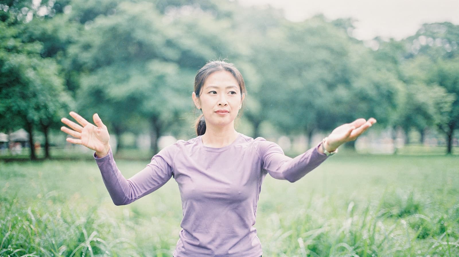 A woman in a purple shirt practices tai chi in a grassy park with trees in the background.