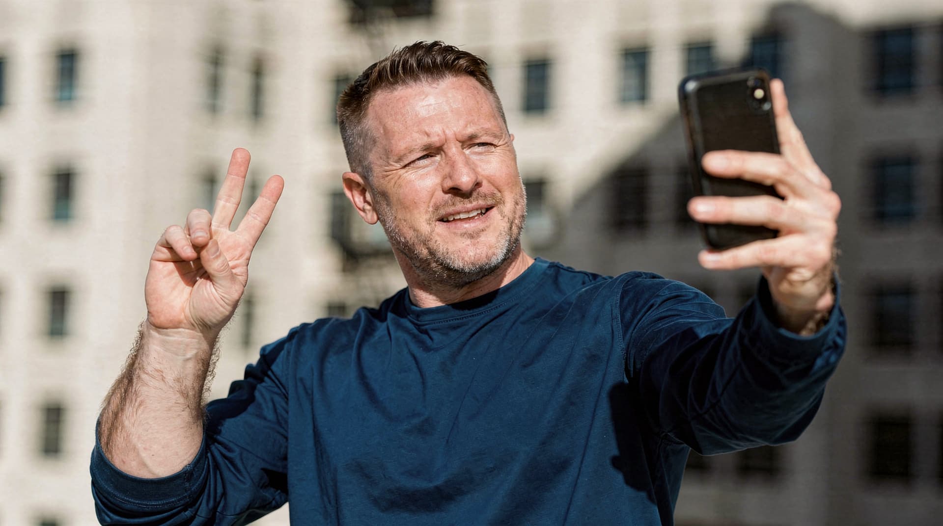 A man in a blue shirt takes a selfie while making a peace sign with his fingers in an urban setting.