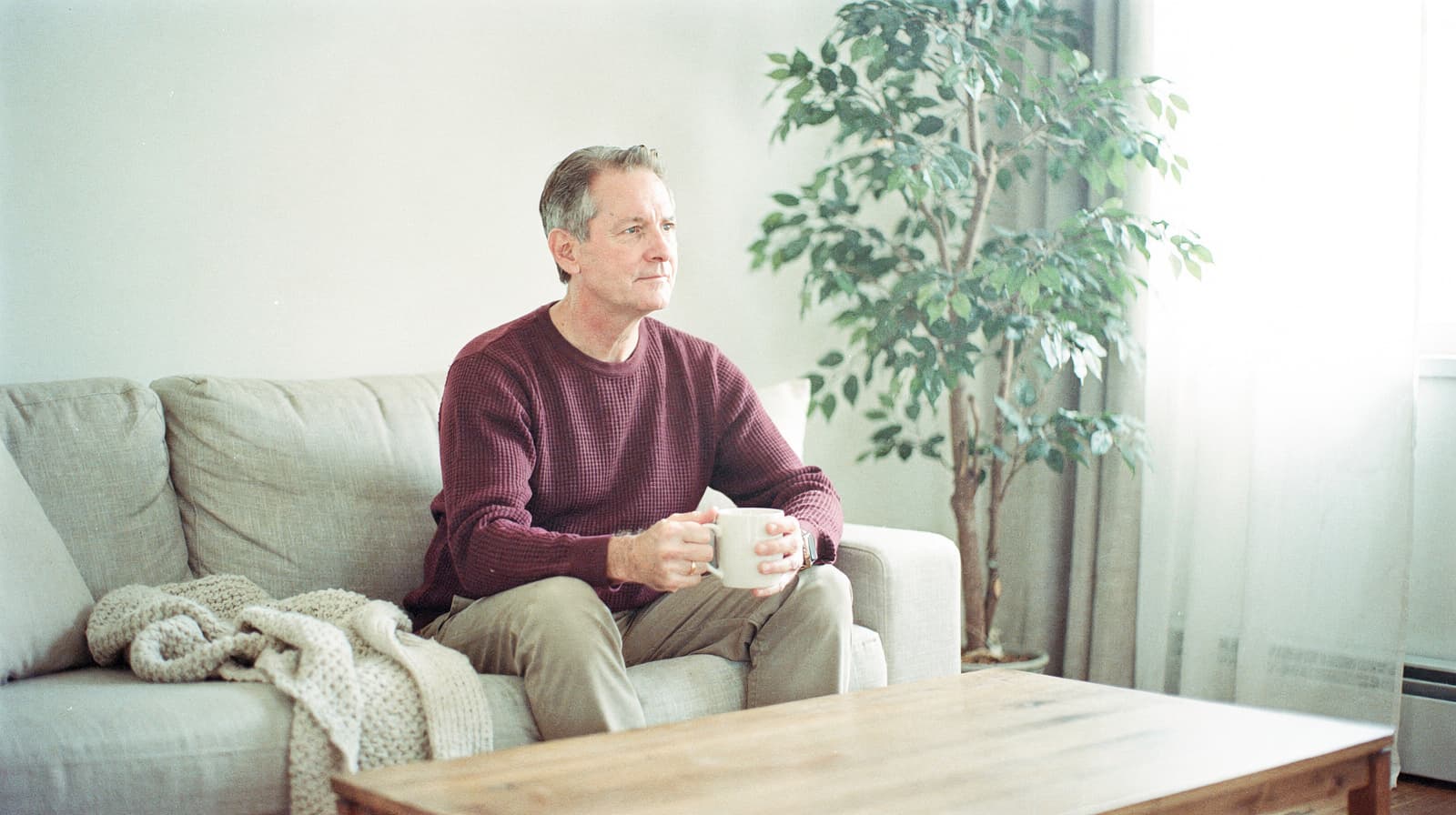 A man sits on a sofa holding a mug, with a plant and a window in the background.