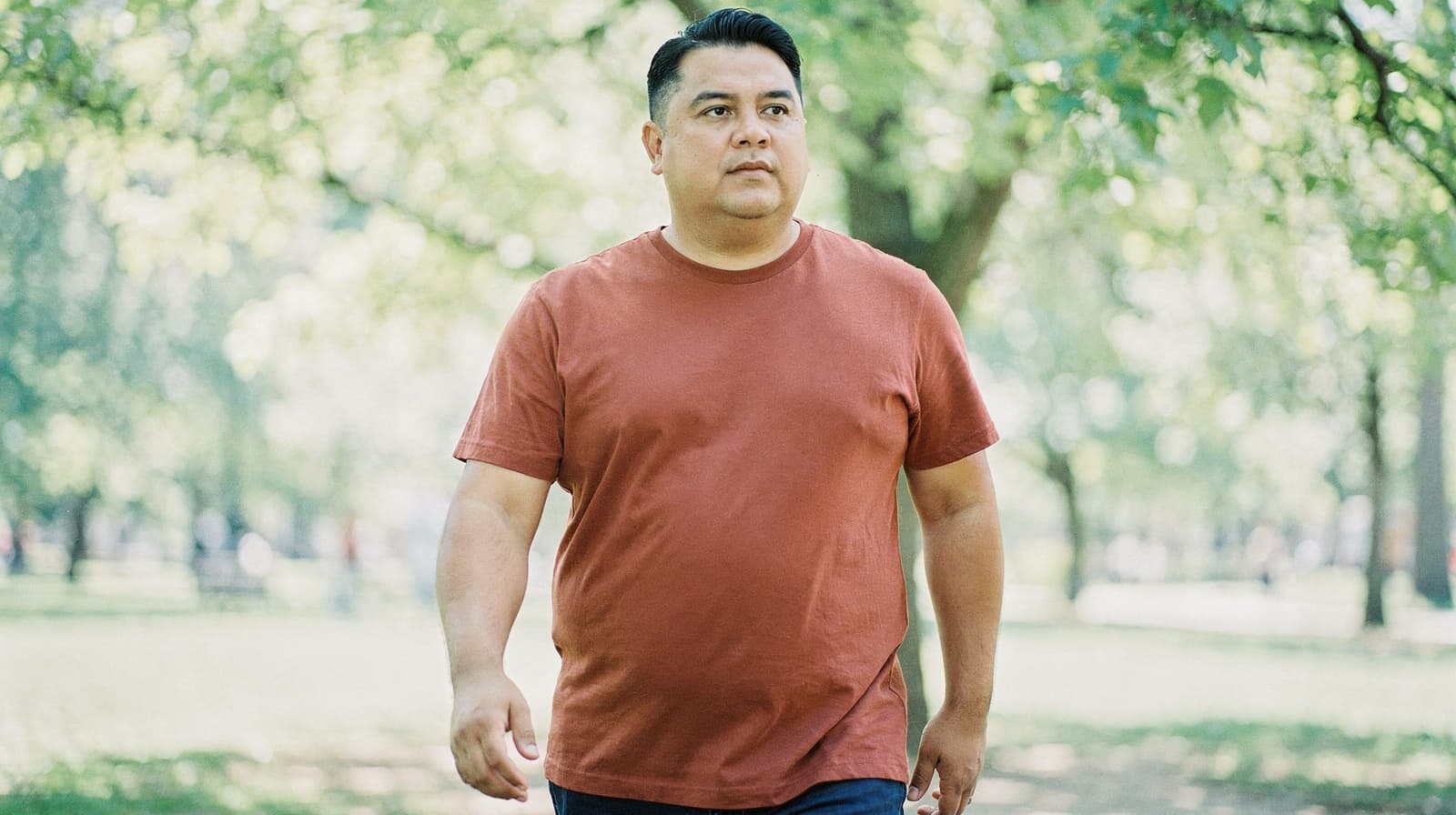 A man in a red shirt walks through a sunlit park with trees in the background.
