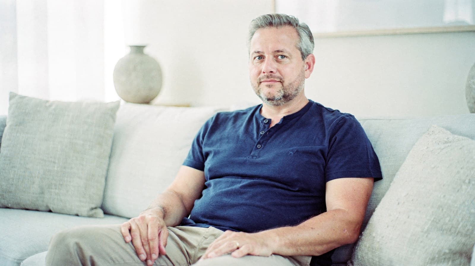 A man in a blue shirt sits on a light-colored sofa in a room with neutral decor.