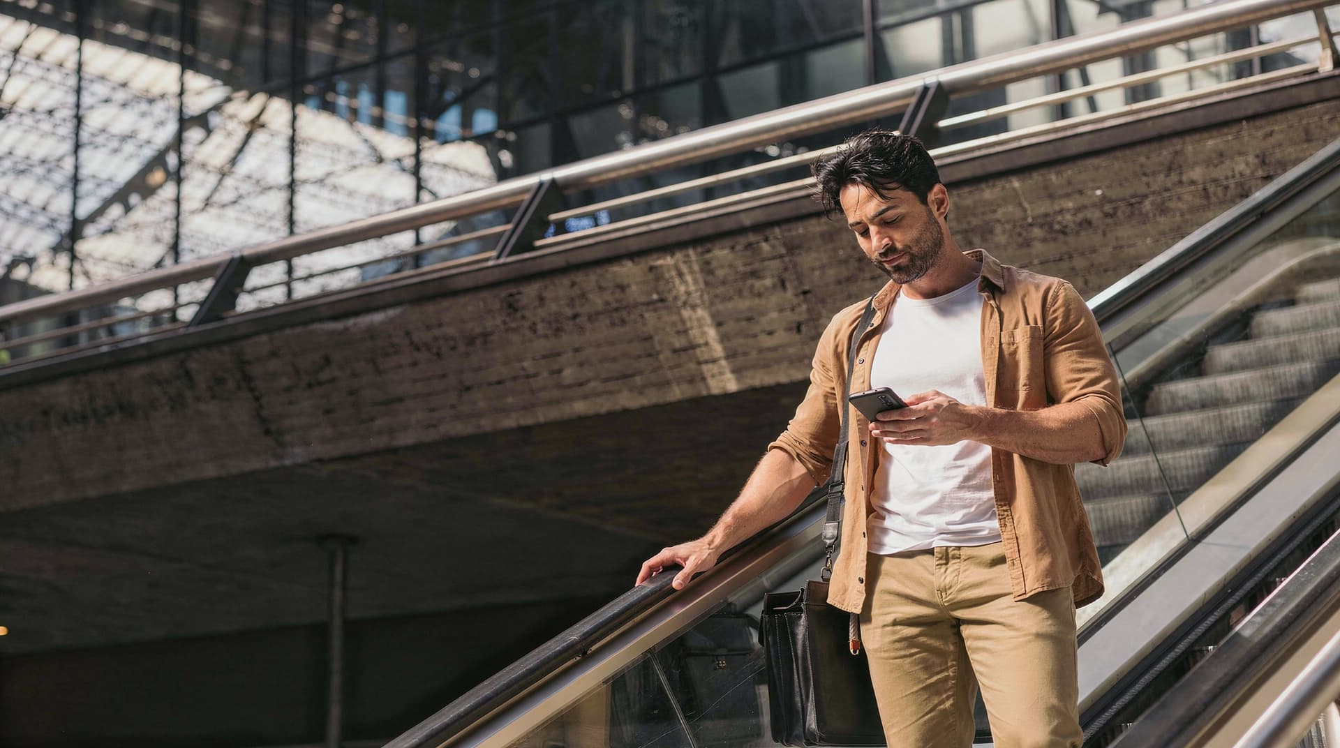 A man in casual clothing stands on an escalator, looking at his phone, in a modern building with glass and metal.