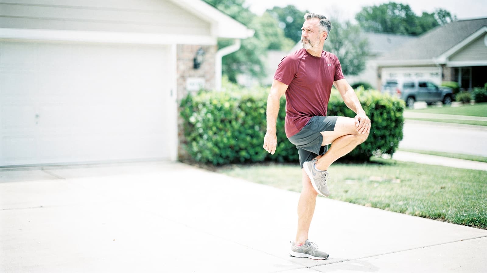 A man in athletic clothing stretches his leg on a driveway in a suburban neighborhood.