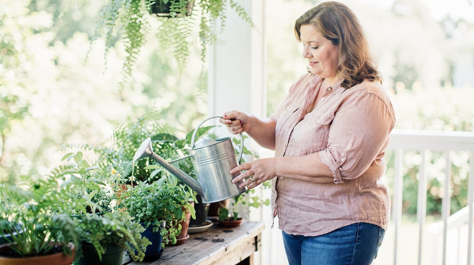 A woman waters potted plants on a wooden table in a sunlit garden.