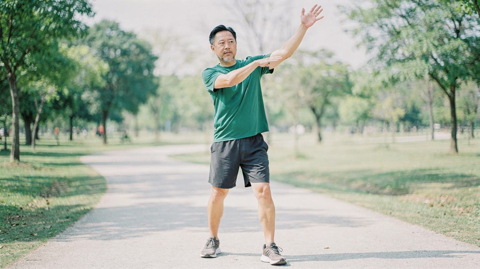 A man in a green shirt and shorts exercises on a park path surrounded by trees.