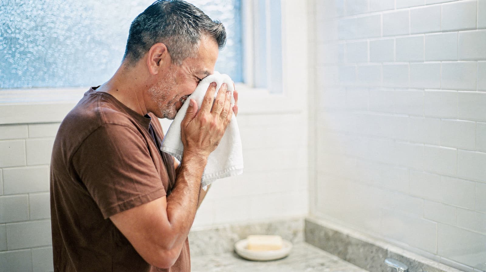 A man in a brown shirt wipes his face with a towel in a bathroom with white tiled walls.