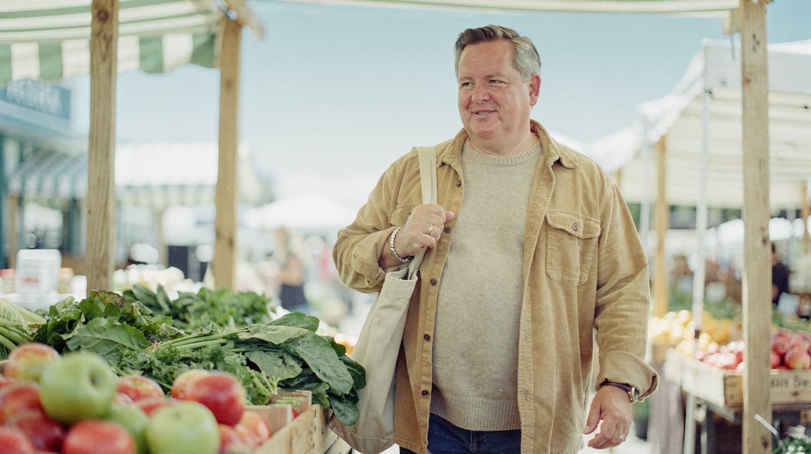 A man carrying a tote bag walks past fruit and vegetable stalls at an outdoor market.