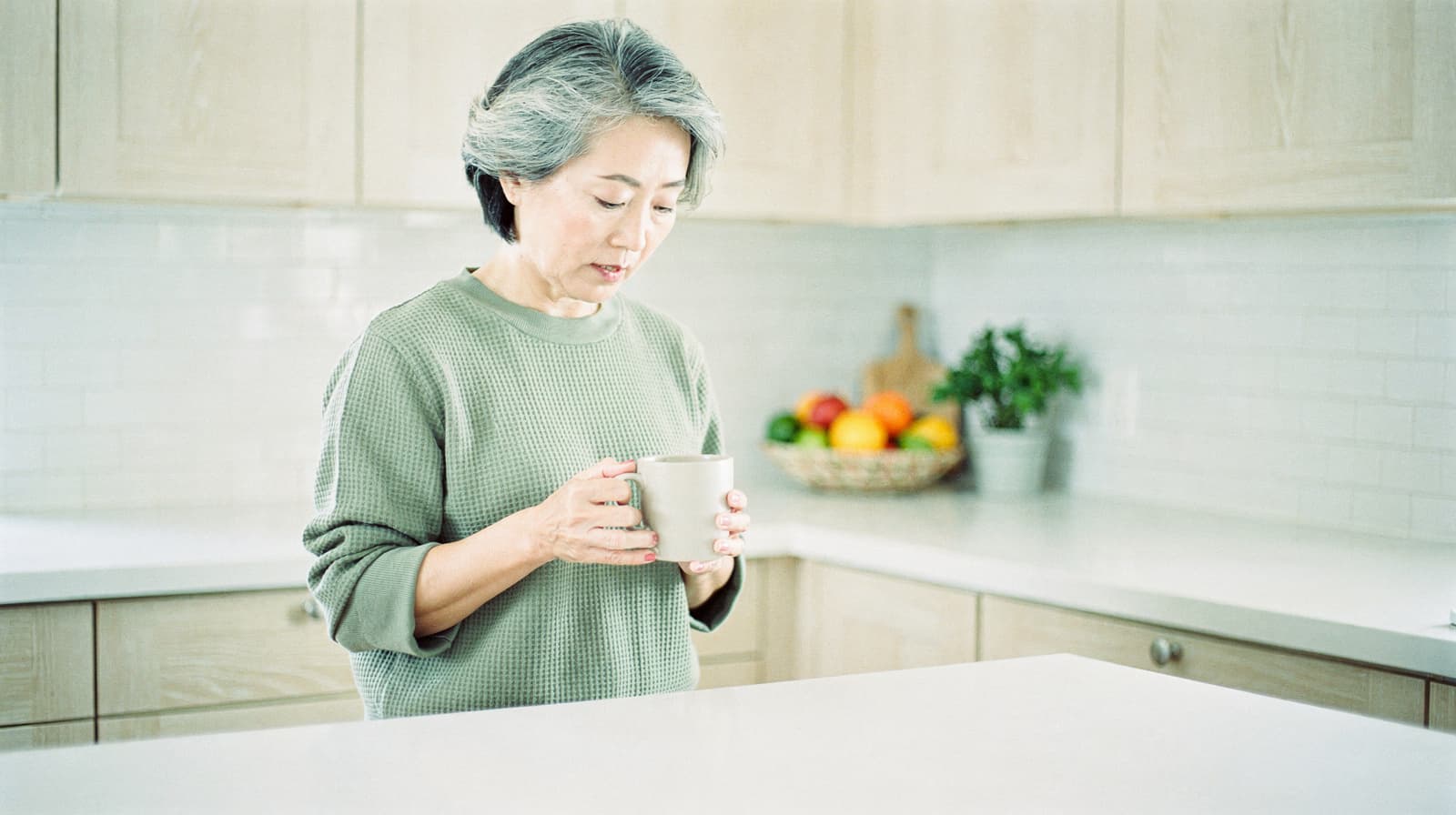 A woman holding a mug stands in a bright kitchen with a fruit basket on the counter.