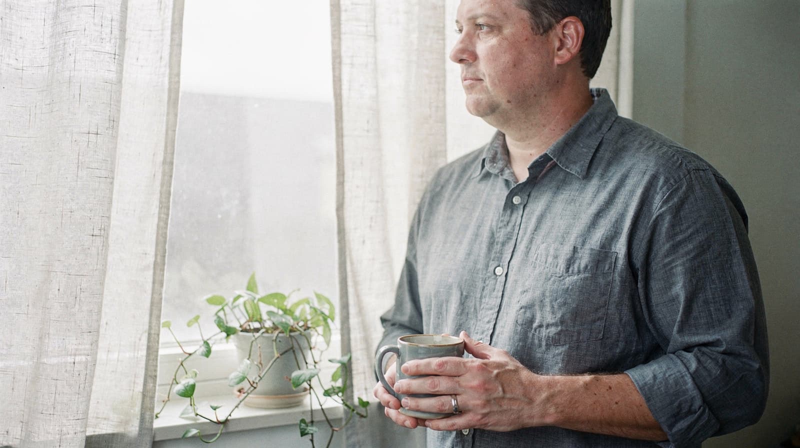 A man in a gray shirt holds a mug and looks out a window with sheer curtains and a potted plant on the sill.