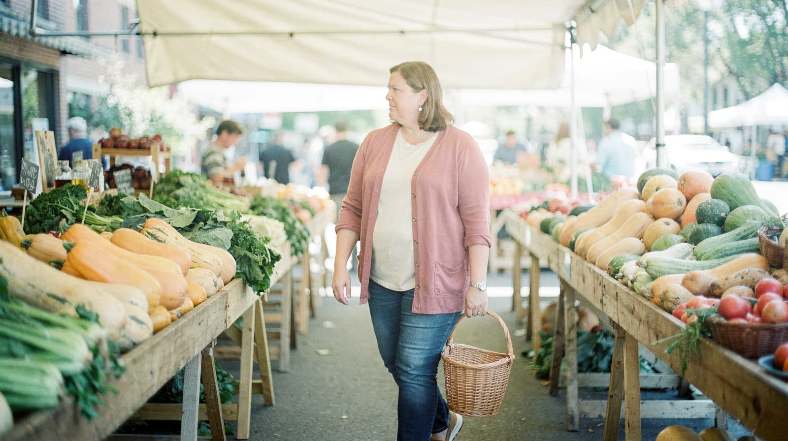 A woman carries a basket while walking through a farmers market with tables of vegetables on either side.