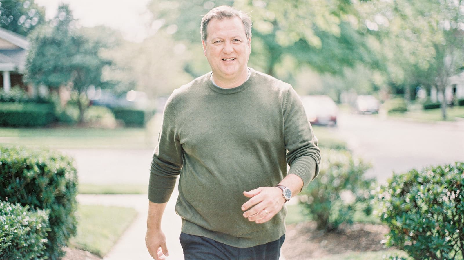 A man in a green sweater walks along a sunny residential street lined with trees and bushes.