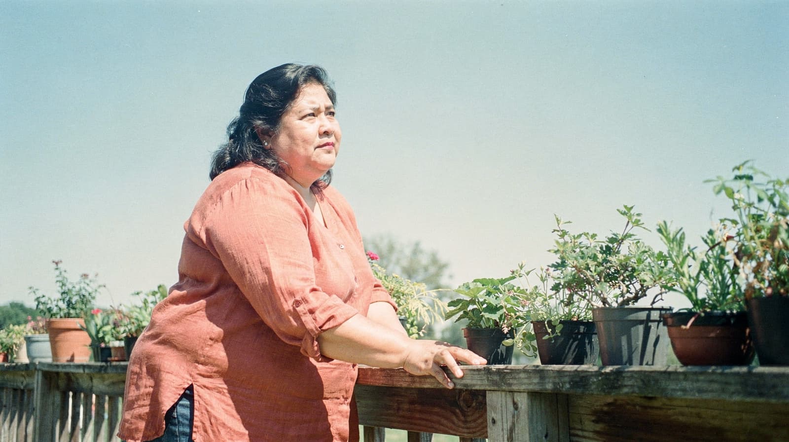 A woman in an orange shirt leans on a wooden railing with potted plants under a clear sky.