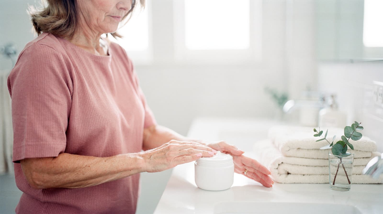 A woman in a pink shirt applies cream from a jar in a bright bathroom with folded towels and a small plant nearby.