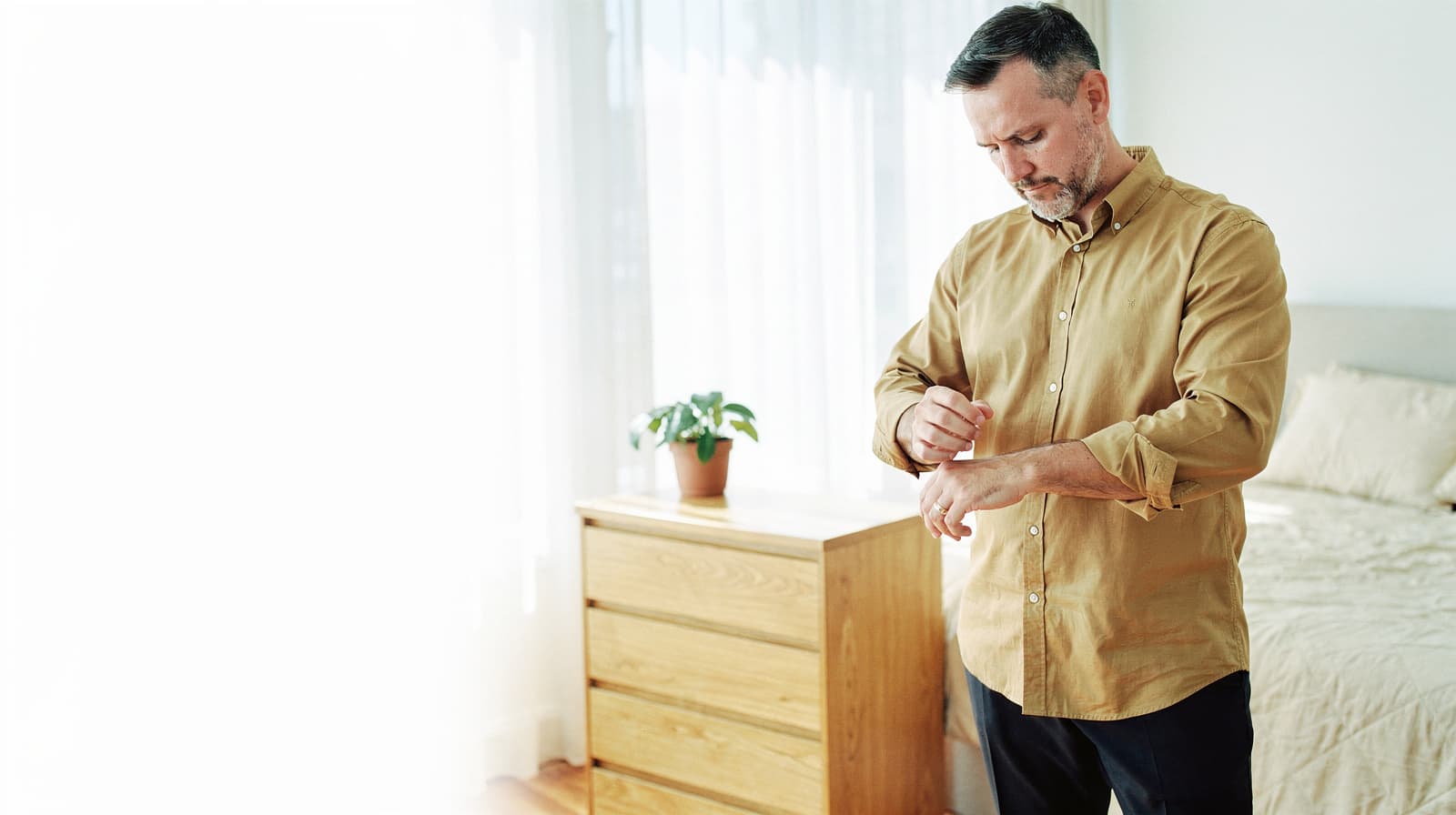 A man in a beige shirt stands in a bedroom adjusting his watch near a wooden dresser with a potted plant.