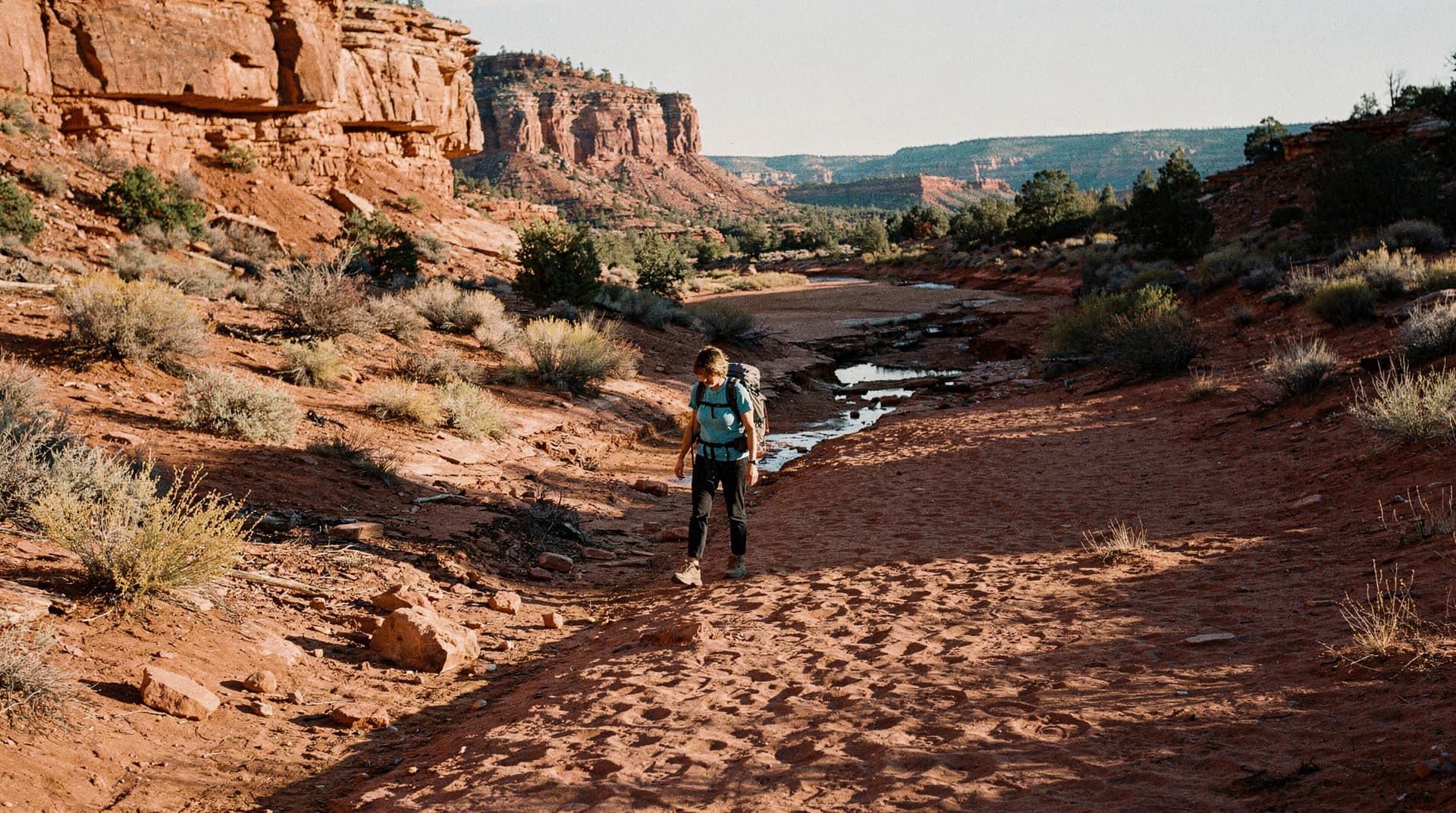 A person with a backpack walks along a sandy trail in a desert canyon landscape with red rock formations.