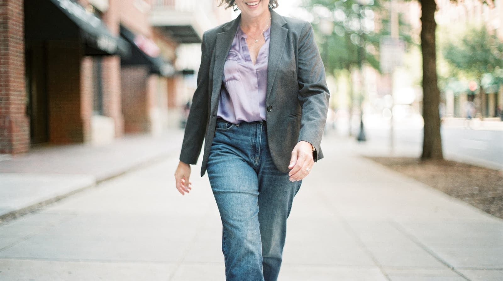 A woman in a blazer and jeans walks confidently down a city sidewalk.
