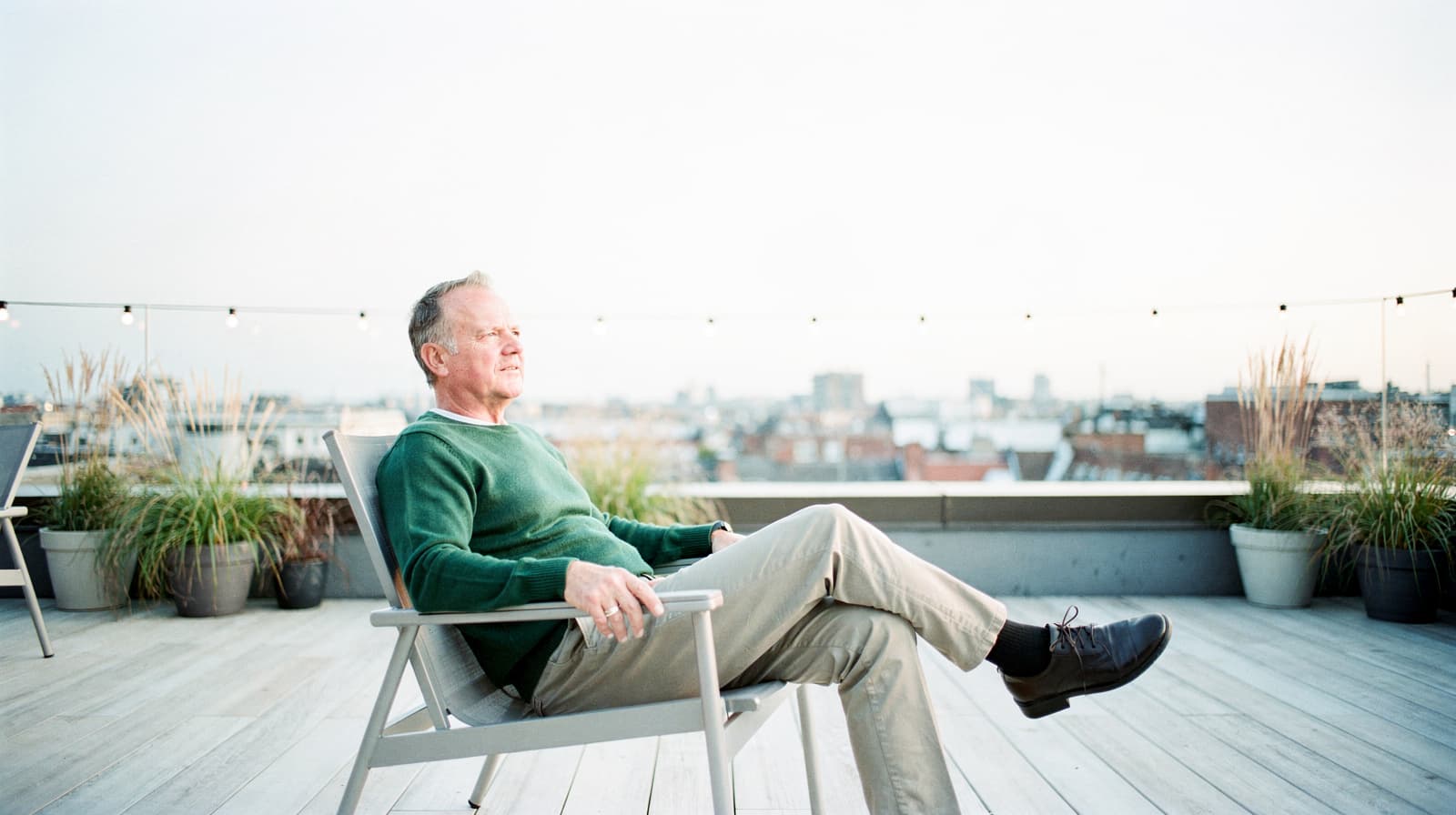 A man in a green sweater sits on a rooftop patio chair, surrounded by potted plants, with a cityscape in the background.